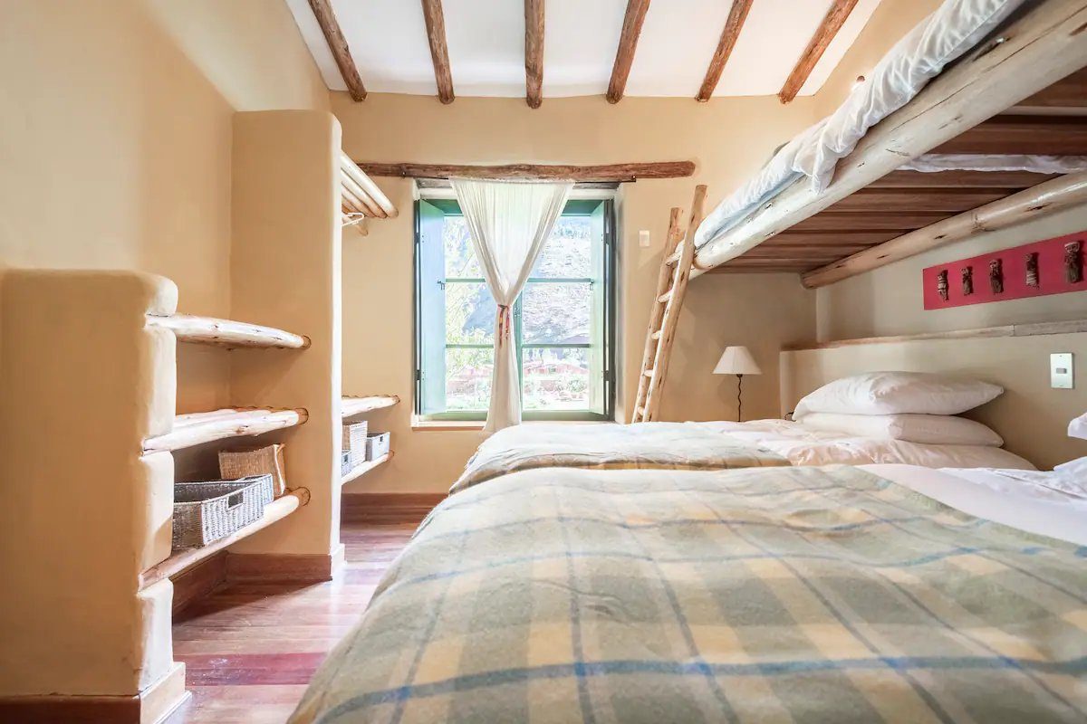 A bedroom under a beamed ceiling with a double bed, a bunk above it reached by a wooden ladder, a checked blanket and a green framed window