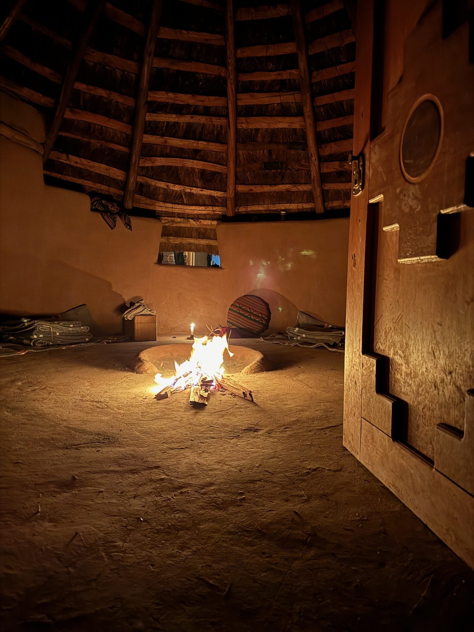 A fire burning low in the middle of a round adobe room at night, under a roof of wooden beams, with folded blankets along the wall and a candle beside the hearth