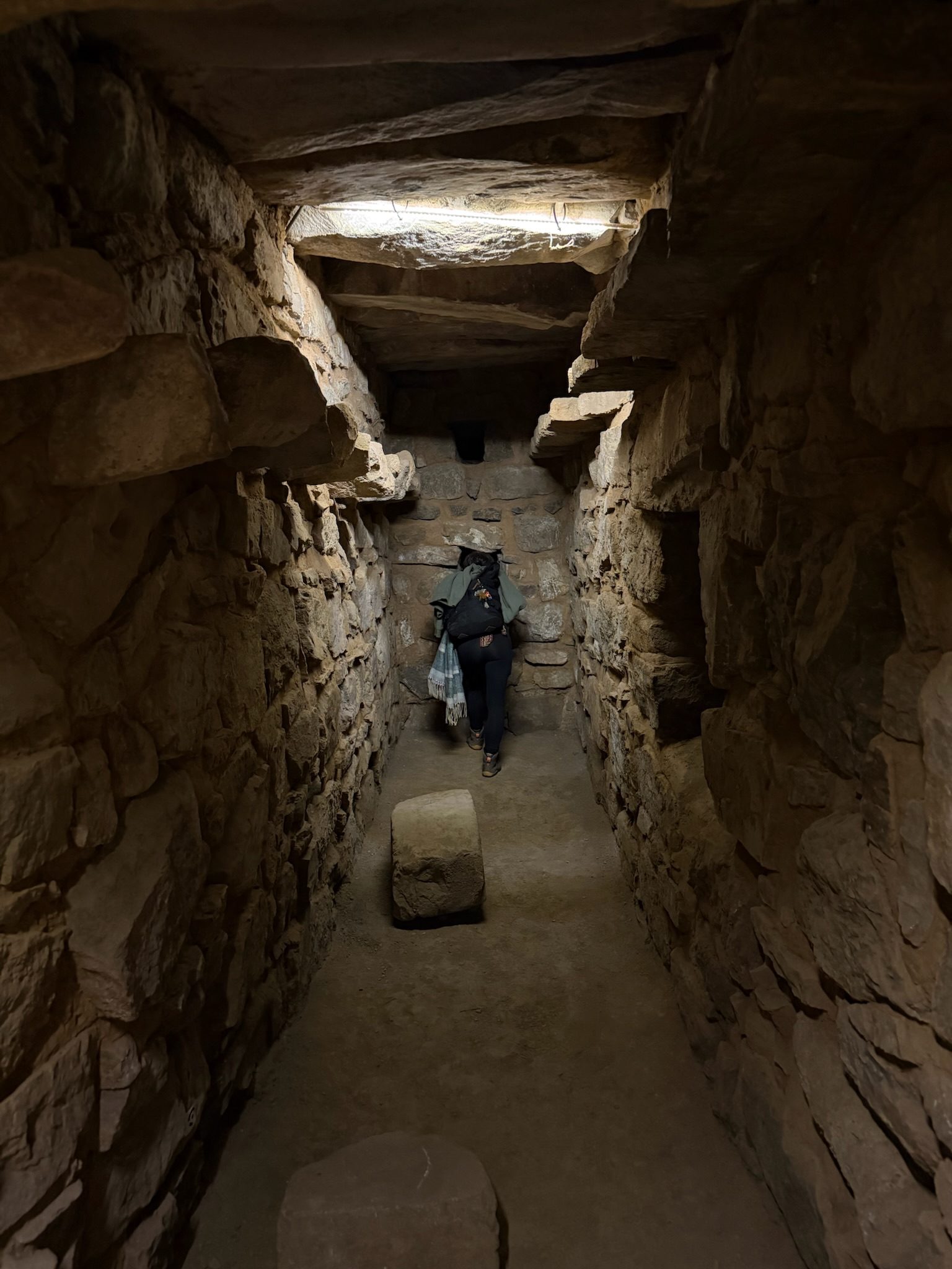 A narrow stone passage roofed with heavy slabs, lit by one lamp, a person standing at the far end