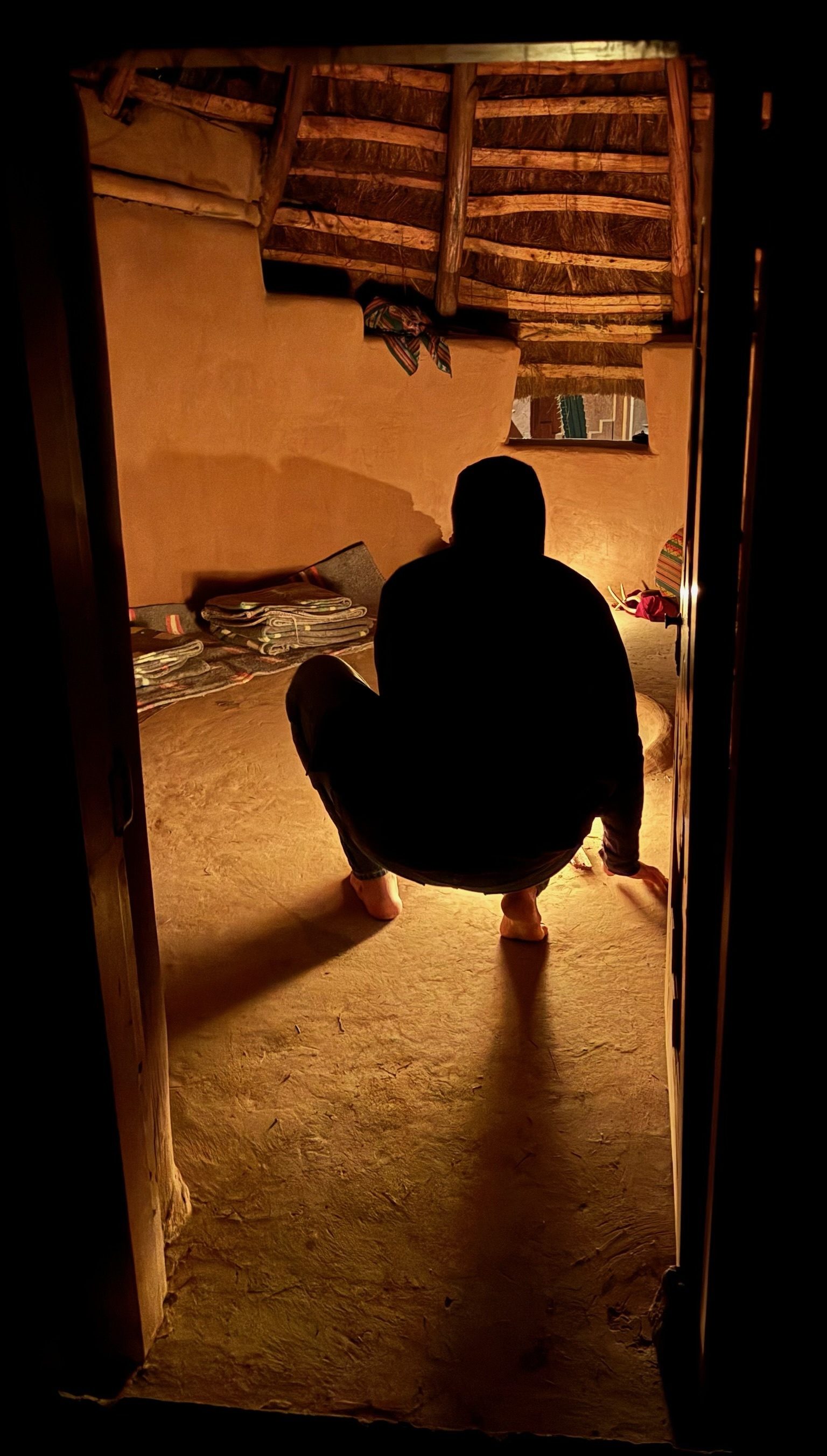 Someone crouching barefoot on the earth floor of a round room lit by firelight, seen from behind through the open door