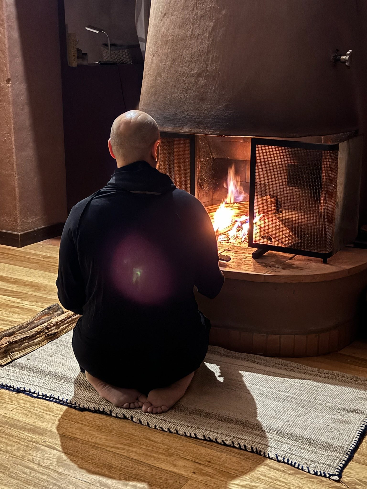 Someone kneeling barefoot on a rug before a fire burning in a round clay hearth, seen from behind