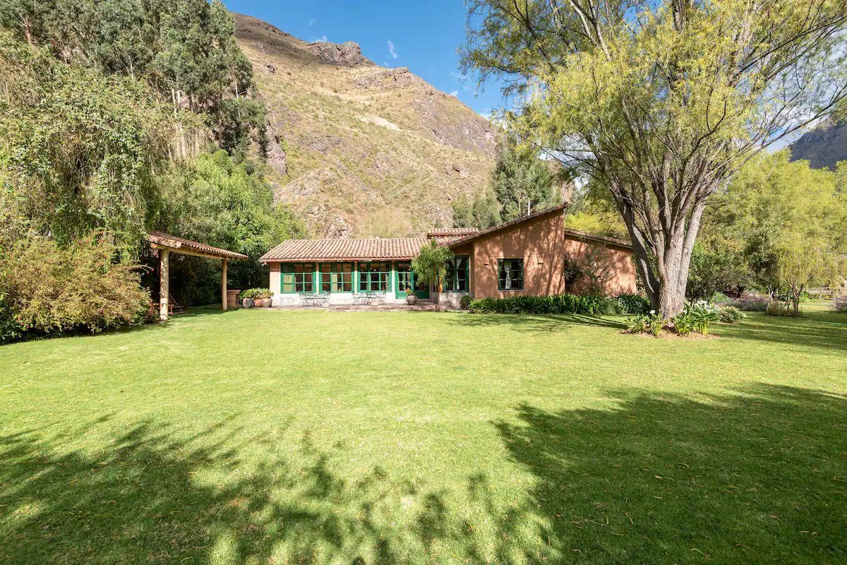 An adobe house with a tiled roof and green framed windows across a wide lawn, a covered terrace at one end and a mountain rising behind it
