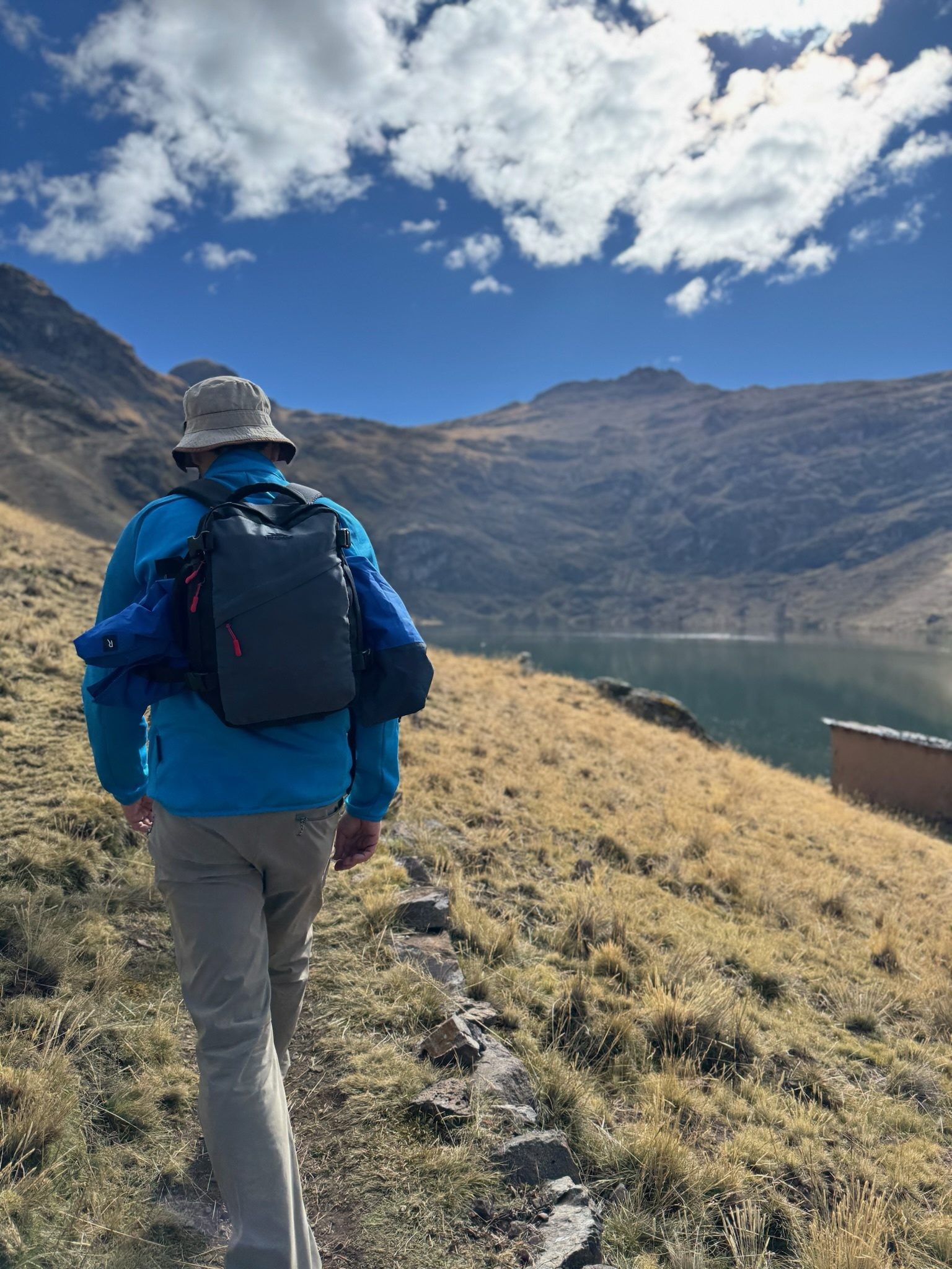 A walker with a rucksack and a sun hat on a stone edged path above a high lake, mountains and cloud ahead
