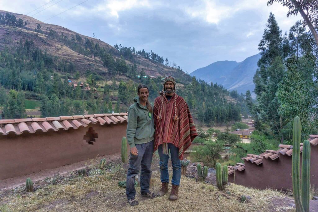 Two people standing together outside an adobe house, with cacti and mountains behind them