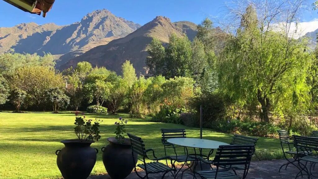 A stone terrace set with metal garden chairs and two planted urns, looking across a lawn and trees to a bare mountain